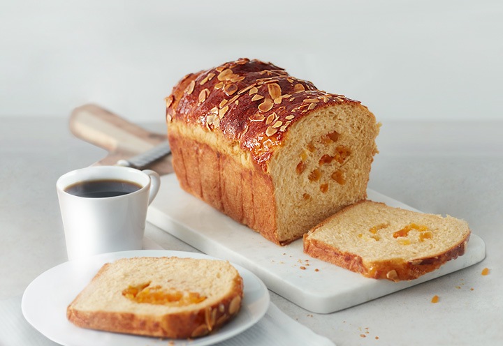Um pão de brioche de alperce seco com amêndoas laminadas está numa tábua branca.