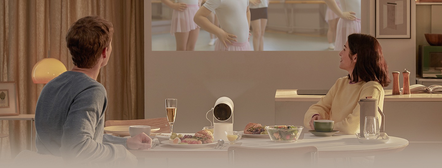 Um casal desfruta de uma refeição. Colocado na mesa de jantar, The Freestyle projeta uma cena de uma aula de ballet infantil na parede.