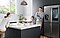A mother and daughter eat salad with orange juice in the kitchen, while the father watches over them and smiles while standing next to the Family Hub, where the screen display indicates rainy weather and reads "Good Morning Chris."