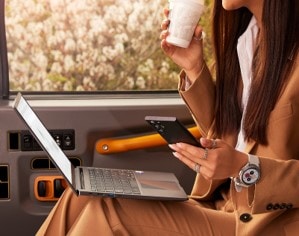Femeie într-un vehicul, ținând o cafea, un smartphone și un laptop