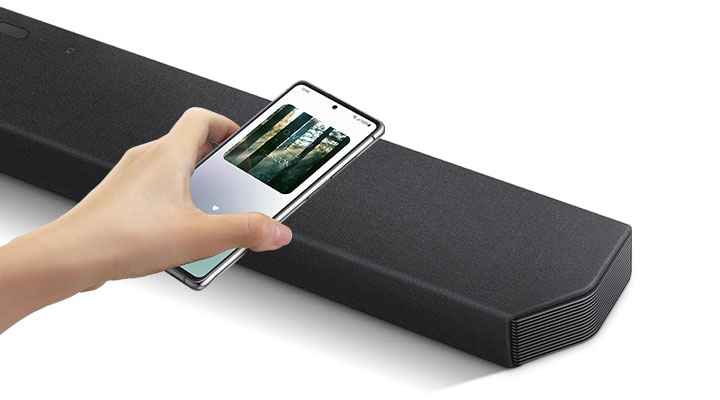 O mână atinge un smartphone, cu aplicația pentru muzică Samsung pe ecran, de soundbar.