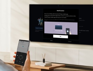 Într-o sufragerie elegantă, o persoană folosește un telefon Galaxy pentru a controla un televizor Smart Samsung prin telecomanda virtuală a aplicației SmartThings, vizibilă pe ecranul telefonului, cu televizorul vizibil în fundal