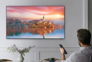 Într-o sufragerie elegantă, un bărbat stă pe canapea și folosește telecomanda inteligentă Samsung pentru a naviga pe televizorul Smart Samsung, care prezintă o imagine cu un peisaj