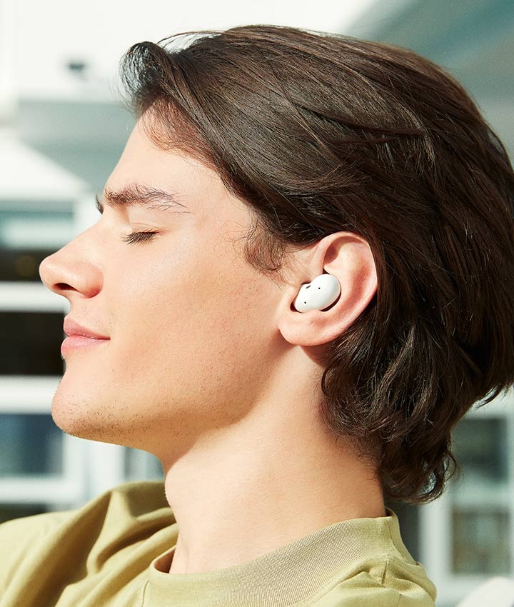 Snimak profila čoveka koji se izležava na suncu na terasi i sluša muziku uz svoje Galaxy Buds Live slušalice.