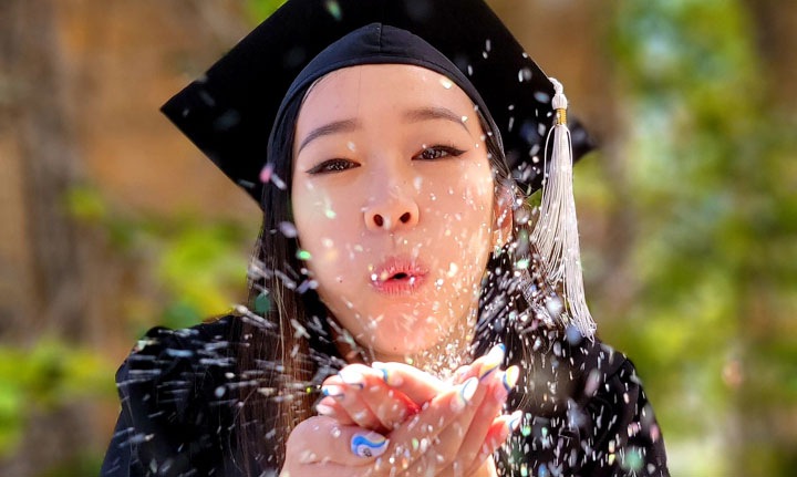 Džesika duva konfete sa svog dlana gledajući u kameru. Nosi crnu diplomsku kapu i mantil na otvorenom prostoru. Pozadina je zamućena da bi se postigao bokeh efekat.