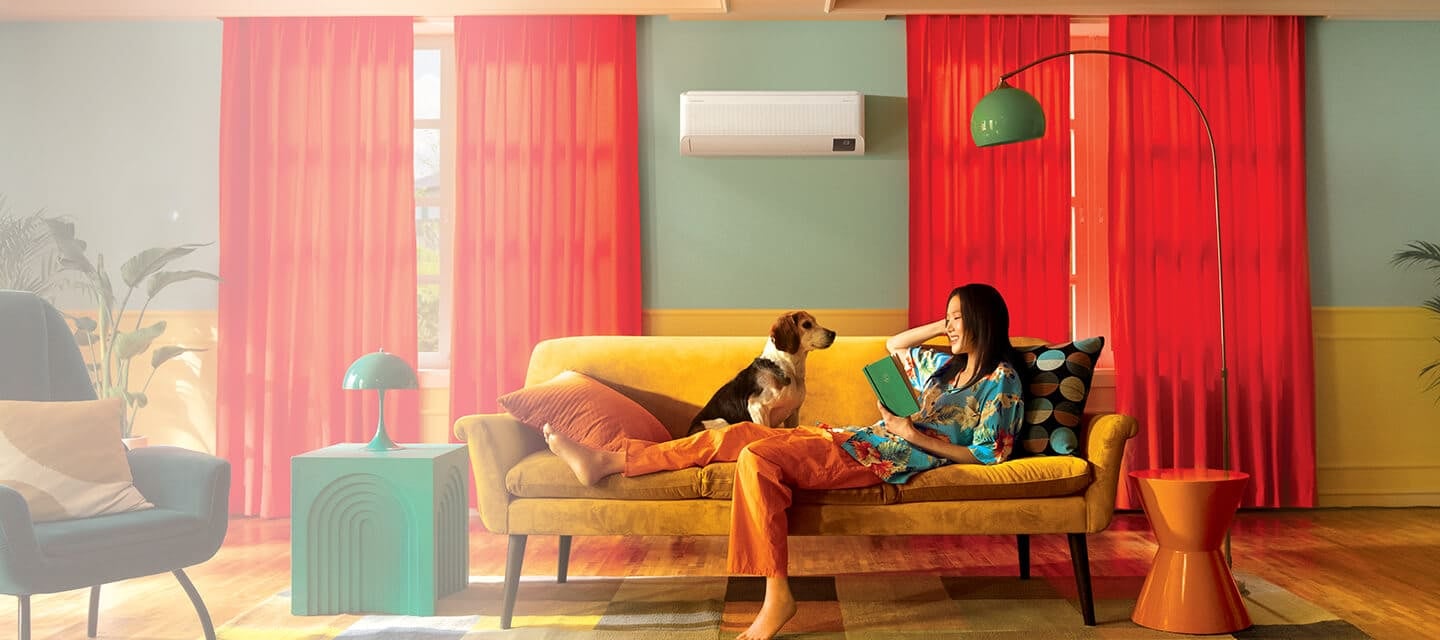 Девушка с собакой отдыхает на диване в середине комнаты, держа книгу в руке, под кондиционером Samsung с технологией WindFree