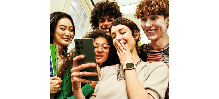Группа друзей, сидя снаружи, смеясь и глядя на свои смартфоны. Там показаны значок Galaxy Store и другие значки.