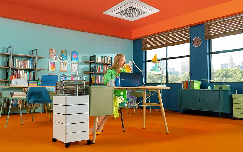 Женщина сидит под 4-сторонней кассетой в стильном и красочном офисе. На ее столе раскрыта черная книга Galaxy Book.