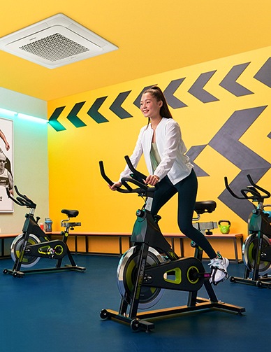 Женщина занимается на велотренажере под 4-сторонней кассетой в тренажерном зале с желтыми стенами.