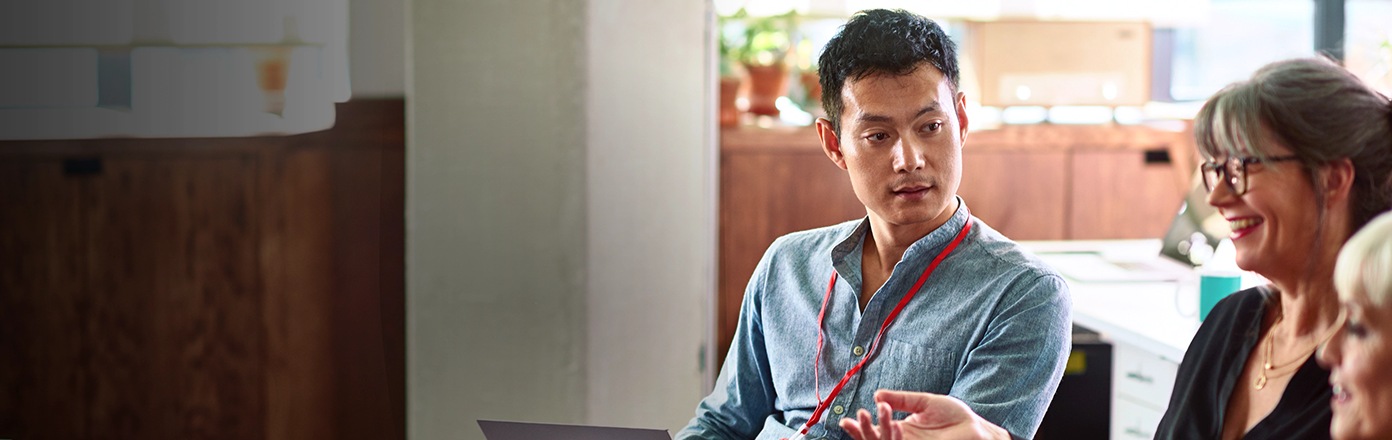 На фото мужчина сидит и, держа серебристый ноутбук на коленях, смотрит на женщину слева, которая изображена не в фокусе.