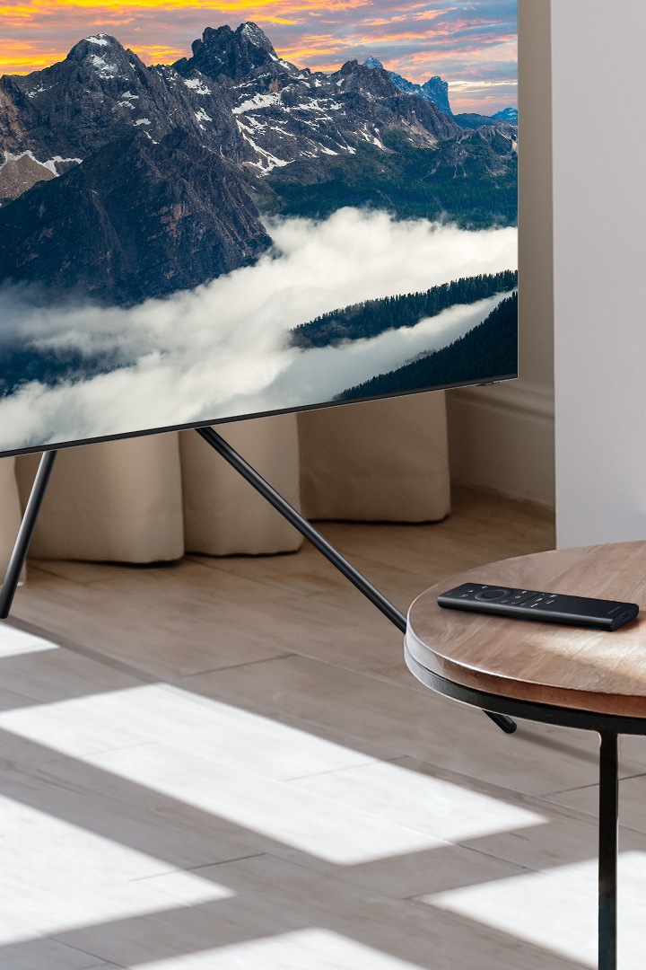 جهاز التحكم عن بعد SolarCell موجود على طاولة صغيرة بجوار كوب. تلفزيون QLED جديد مُثبَّت على حامل تلفاز.