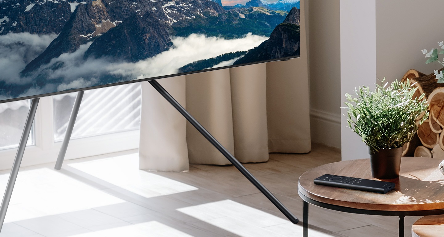 جهاز التحكم عن بعد SolarCell موجود على طاولة صغيرة بجوار كوب. تلفزيون QLED جديد مُثبَّت على حامل تلفاز.