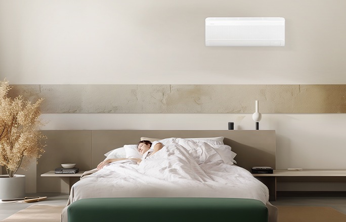 A person sleeps peacefully in a bedroom, lying under a Samsung windfree wall-mounted air conditioner.