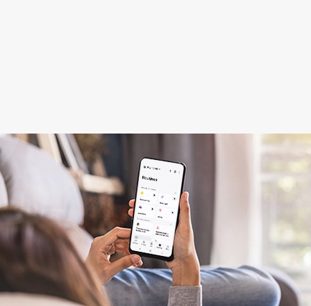 A person relaxes on a couch while holding a smartphone with the SmartThings app open.