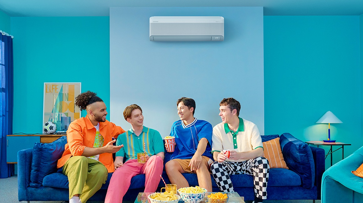 Four young men with colorful attire chat and enjoy snacks on a couch. A WindFree AC is mounted on a light blue wall.