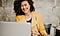 A curly-haired woman laughs while video chatting on her Galaxy laptop. She is wearing a mustard yellow jacket and holding a pen in her right hand. She is sitting in front of a beige cemented wall.