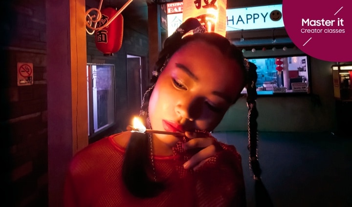 En kvinna iakttar en lång, brinnande tändsticka på en neonmarknad. Master it. Creator classes.