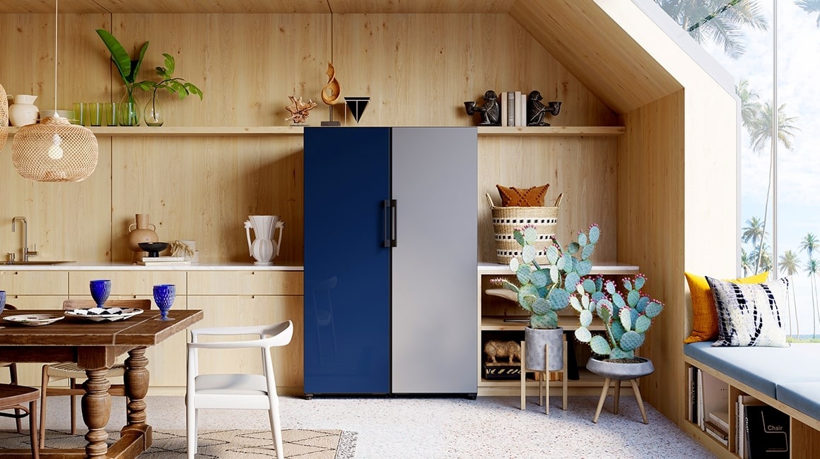 Two 1-door Bespoke refrigerators are placed next to each other in a stylish dining room with plants. The color of the left one is a Glam Navy and the right one is Satin Gray. The handles are placed in the center so the doors open outwards.