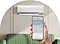The WindFree AC is mounted on a white wall behind a green sofa. A hand holds a smartphone, displaying the “Room Air Conditioner” screen.