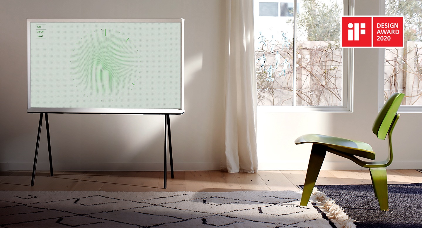 A Cloud White model of The Serif sits on a black stand in a bright and elegantly simple living room, with an area rug, a stylish three-legged green chair, and large windows. The image on screen is Leaf Ambient Mode with clock ticking.