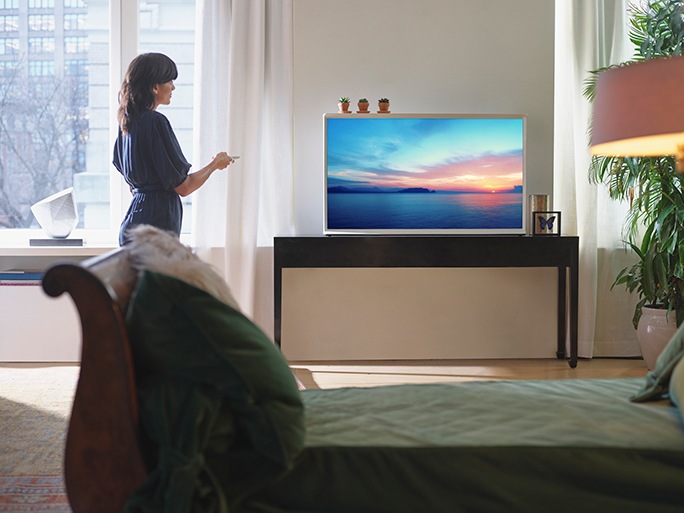 A woman sits on her bed tying her shoelaces. A Cotton Blue model of The Serif rests neatly on a TV stand in the corner with time and weather information on the screen using Ambient mode.