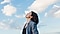 A woman in a blue-gray blazer stands with her head tilted toward a blue sky with white puffy clouds. Her eyes are closed and she looks to be inhaling deeply.