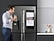 A man drinks a glass of water in the kitchen while checking on his sleeping daughter from the Family Hub screen connected to SmartThings.