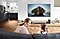 A mother and daughter watch a movie on the big screen TV in the living room, while the father is seen in the background grabbing snacks from the kitchen and watching the same movie through the mirrored feed on the Family Hub screen.