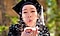 Jessica blows confetti from the palm of her hand while looking at the camera. She is wearing a black graduation cap and gown outdoors. The background is blurred for a bokeh effect.
