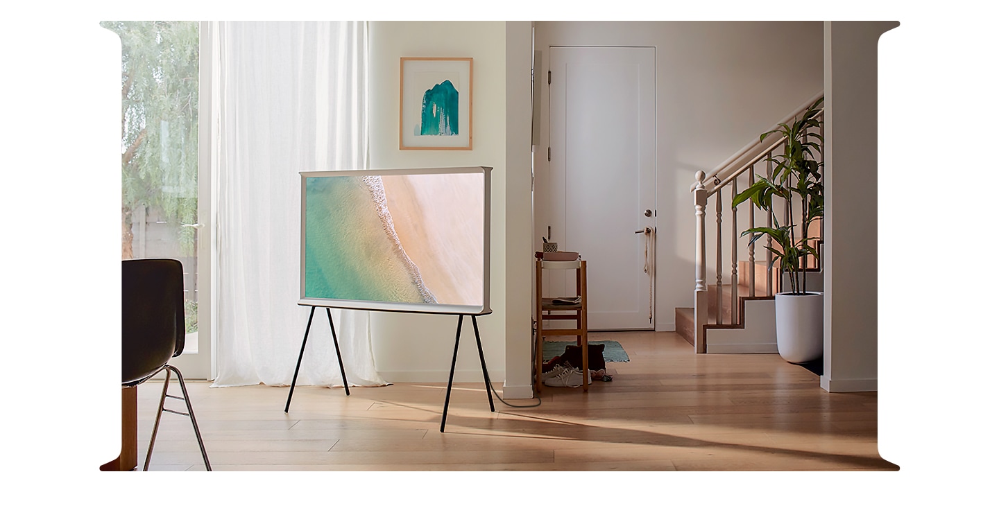 The Serif from 4 different perspectives demonstrate how amazing it can look from all different angles. In the first scene, we see a Cloud White model of The Serif on a black stand in a simple and elegantly styled room with the onscreen image being a bird’s eye view of a beach showing a wave as it meets the sand. The scene shifts to a man and a woman having fun preparing food on a kitchen island. A Cloud White model of The Serif is resting directly on the kitchen island in front of them, showing the Fabric pattern in Ambient Mode in a salmon color. The scene shifts again, and this time the back of a Cloud White model of The Serif is shown on a black stand in the foreground of an artist’s workshop. In the background to the right, a man is sitting next to a long worktable, playing with a dog. In the final scene, a Cloud White model of The Serif is on a black stand in the middle of a bright and airy room, with its back to the window. The onscreen image is a beach showing a large wave meeting the sand and a surfer on his board paddling out to the ocean.