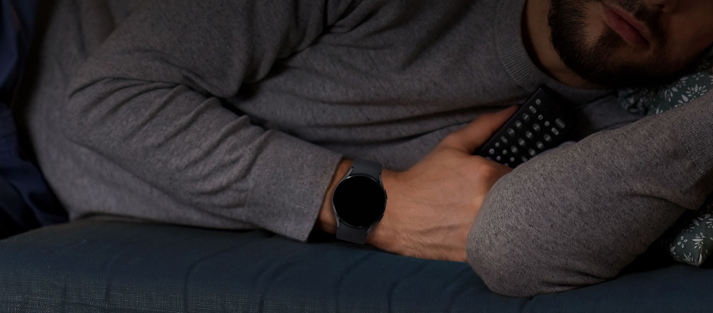 Moški, ki nosi Galaxy Watch uro, spi, v roki pa drži daljinski upravljalnik.