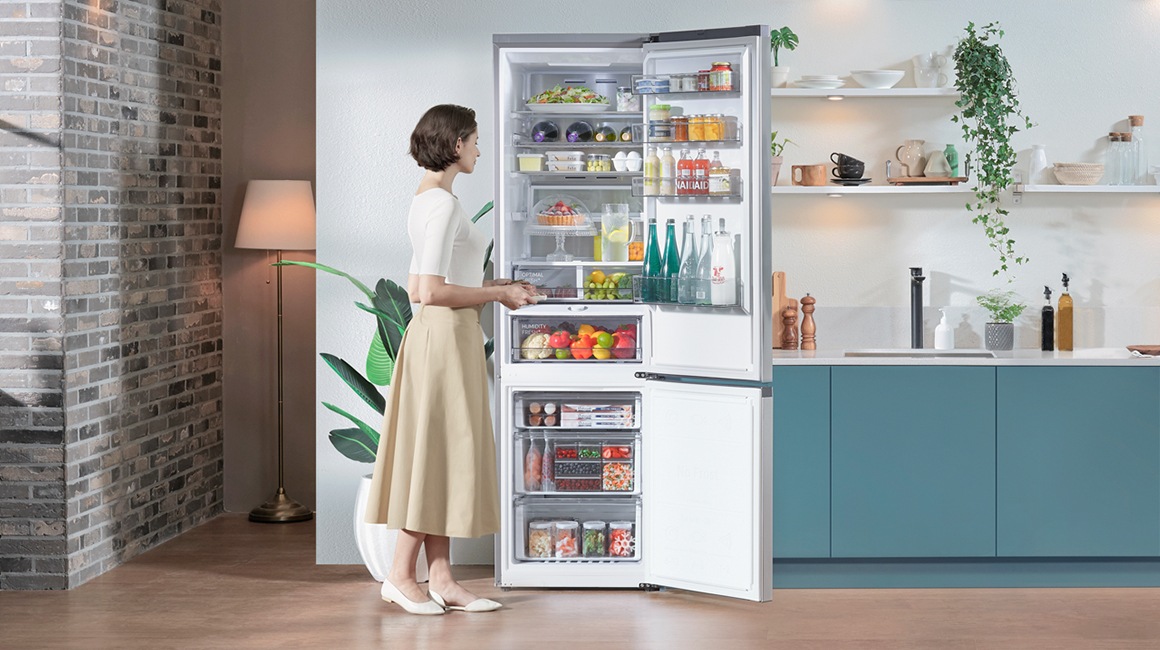 V sodobni minimalistični kuhinji s sivomodrimi pulti ženska odpira hladilnik Samsung, ki je hladilnik z veliko prostornino, znotraj pa je veliko lepo razporejenih živil.