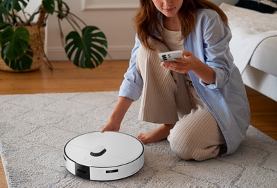 A woman kneels on a rug, using her phone to control a white Samsung Jet Bot Combo in a tidy living room.