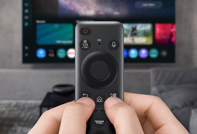 Hands holding a Samsung Smart Remote aimed at a Samsung TV displaying apps.