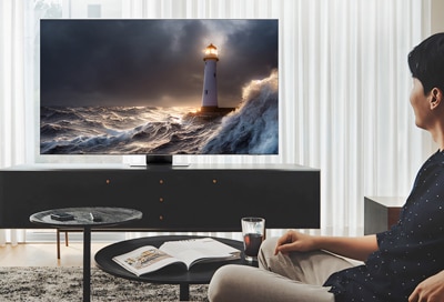 A person sitting in a modern living room, watching a Samsung Smart TV displaying a dramatic scene of a lighthouse amidst stormy seas.