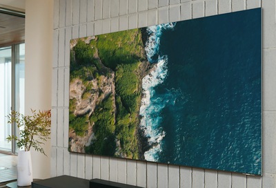 Large wall-mounted Samsung TV displaying a high-resolution aerial view of a rugged coastline, with clear blue waters on the right and green cliffs on the left, enhancing a modern room's decor.