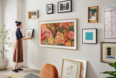 A woman uses a tablet to interact with The Frame TV displaying a vibrant floral painting, surrounded by various framed artworks on a wall.