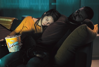 A couple asleep on a couch, the woman resting her head on the man's shoulder, both holding a popcorn bucket, under a dimly lit ambiance.