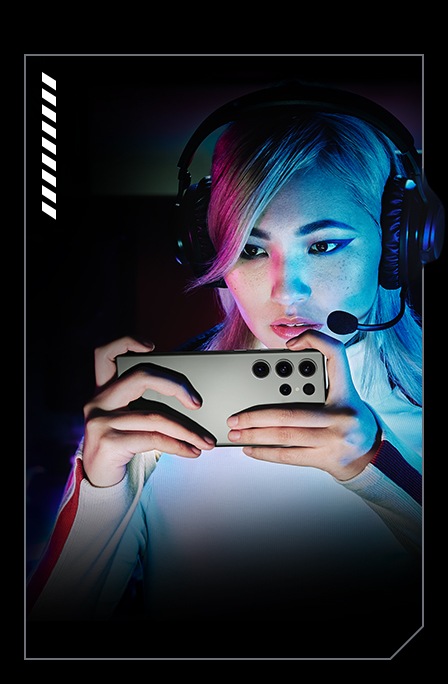 A woman is playing a game on her Galaxy S23 Ultra with a gaming headset on. The background is dark while her face and the Galaxy S23 Ultra in her hands are brightly lit with neon lights.