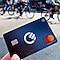 A person holds a dark blue Mastercard credit card with a contactless payment symbol, featuring a circular logo in the center. Blurry bicycles are in the background.