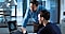 In a cool-tone modern office setting, two men are looking at a Galaxy Book screen and discussing its contents. One man is sitting at a desk and pointing at the screen, while the other is leaning over the desk to see the screen.