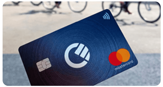 A person holds a dark blue Mastercard credit card with a contactless payment symbol, featuring a circular logo in the center. Blurry bicycles are in the background.