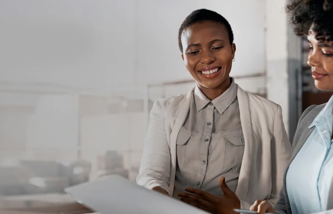 Two women in business attire smile while reviewing a document together.