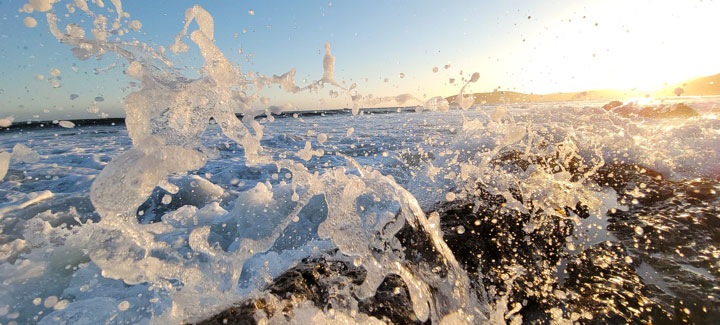 A close up of a wave crashing against rocks in the foreground. Water splashes in front of the camera, lit by the sun hanging low in the sky just off camera.