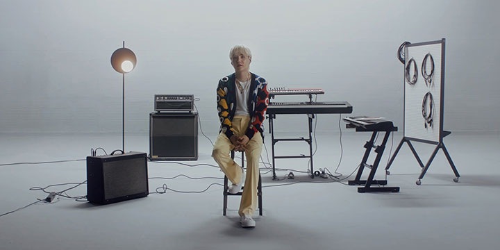 SUGA von BTS sitzt auf einem Hocker, der mitten in einem leeren Tonstudio steht. Er ist von Musikinstrumenten umgeben. Links im Raum steht ein Lautsprecher und rechts stehen drei elektronische Keyboards.