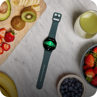 A birds eye view shot of a Samsung Galaxy smartwatch on a counter surrounded by fruit on a chopping board and in bowls.
