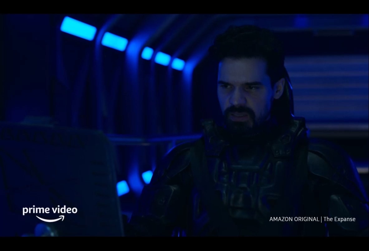 A movie-like content is being shown. A man is facing the camera looking sternly at a screen that is off-camera. Behind him is a dark open space that is illuminated by blue neon lights. Then a spaceship emerges from the right and is seen following a rocket ship which quickly accelerates away, emitting a flash of blue light from its rear. A spaceship then detaches from a metal structure and reverses backwards. In the screen, there are Amazon Prime logo and “Amazon Original | The Expanse” text, as well as a disclaimer saying “Amazon Prime membership or Prime Video fees apply. See primevideo.com/terms for details.