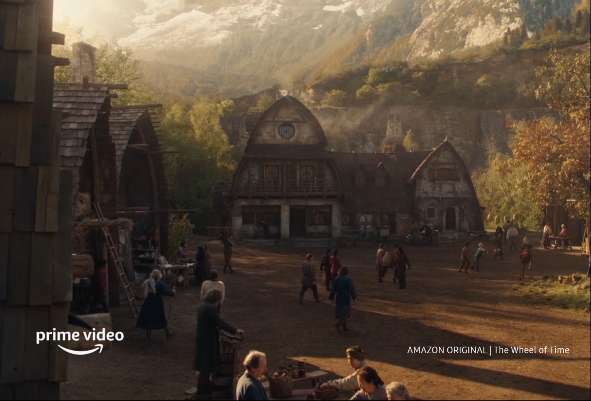 There is a video of a village setting against a mountainous backdrop. People walk through the village in the background and in the foreground, a small group of villagers are sat on a bench sorting through crops. Inside a dimly lit bar, people are seen conversing and drinking beverages from copper cups. Candle lights hang from the ceiling of the room. A lady stands in the doorway of the room in a grey dress with her hands interlocked in front of her body. Behind her are other buildings from the village lit by the sunlight outdoors. A closeup of a young man with red hair shows him smiling at the lady in the doorway. And there are Amazon Prime logo and “Amazon Original | The Wheel of Time” text, as well as a disclaimer saying “Amazon Prime membership or Prime Video fees apply. See primevideo.com/terms for details.”