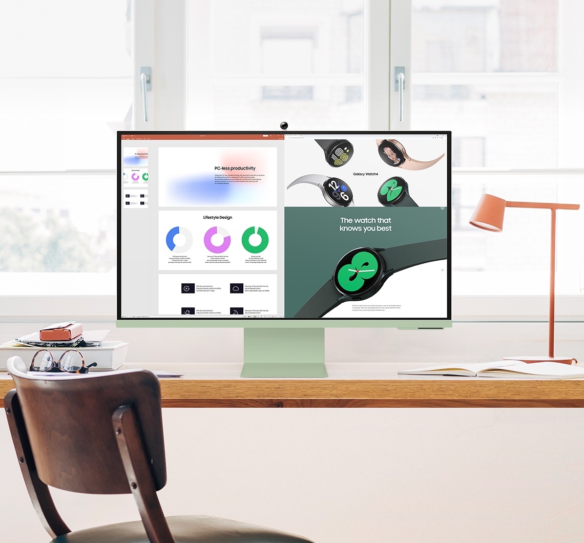 A green Smart Monitor sits on top of a desk. On its screen shows a presentation deck and web page being projected at the same time. A chair is in front of the desk. A desk lamp sits to the right of the monitor.