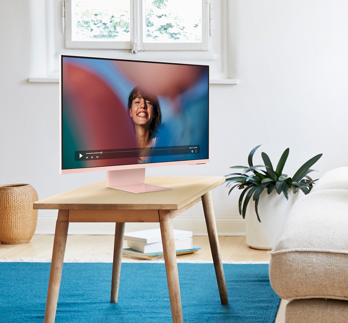 There is a pink Smart Monitor on top of the coffee table in a living room. And a content with a smiling woman is being played on the monitor. A couch and house plant sit next to the monitor.